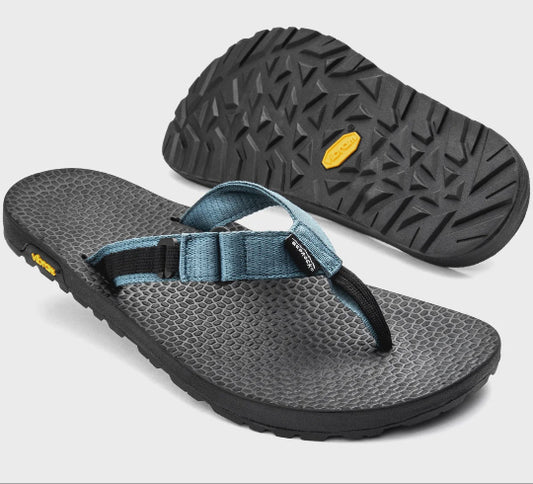A pair of Bedrock brand flip-flops on a light gray background. One sandal is positioned upright to show the textured black outsole with its signature yellow Vibram logo, while the other lies flat to display the gray patterned footbed and blue strap.