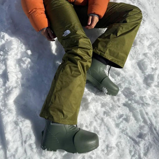 Person wearing green snow boots and pants sitting in the snow with an orange jacket.