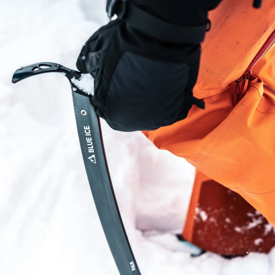 Person wearing orange pants and black gloves holding an ice axe in a snowy environment