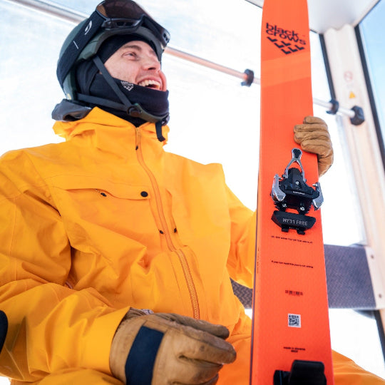 Person in yellow jacket holding an orange ski with a visible Black Crows logo, inside a building.