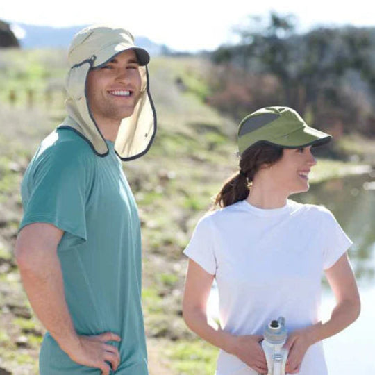 Two people on a lakes edge in sunshine. Man wearing a cream flap cap and woman wearing a green cap.