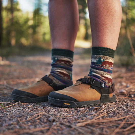 Person wearing patterned socks and brown clogs on a forest floor