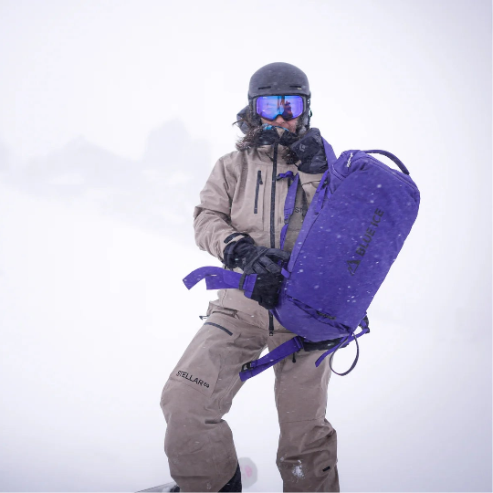 Person in winter gear holding a purple backpack against a snowy background
