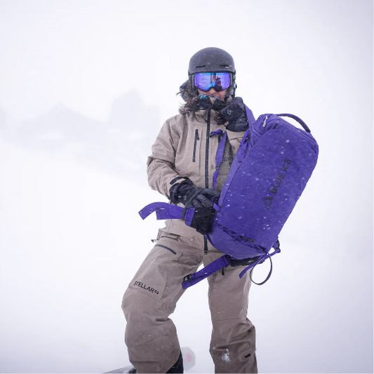 Person in winter gear holding a purple backpack against a snowy background
