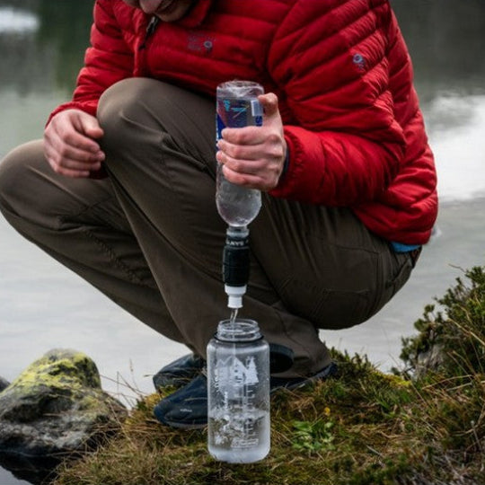 Person in a red jacket using a water filter on a bottle by a lake.